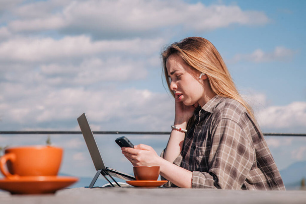 A young, beautiful girl in a plaid shirt talks on a smartphone sitting at an old wooden table in front of a laptop in nature and drinks coffee - Photo, Image