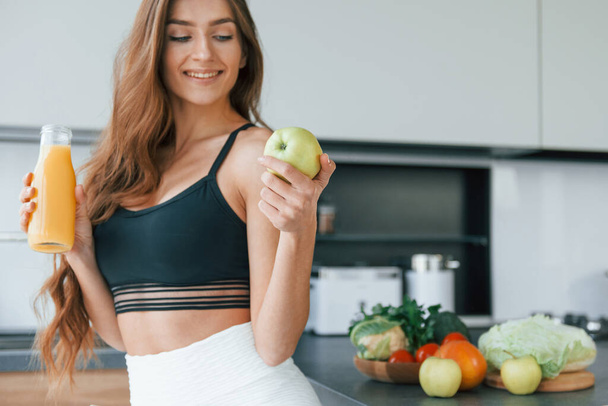 Holds apple in hand. Young european woman is indoors at kitchen indoors with healthy food. - Foto, afbeelding