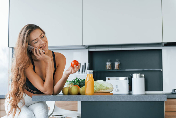 Talks by the phone. Young european woman is indoors at kitchen indoors with healthy food. - Foto, Imagem