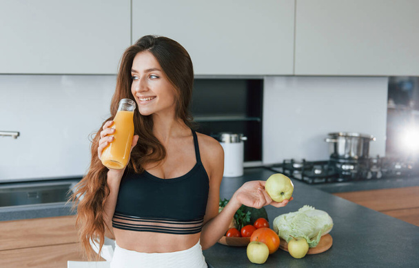 Holds apple in hand. Young european woman is indoors at kitchen indoors with healthy food. - Foto, afbeelding