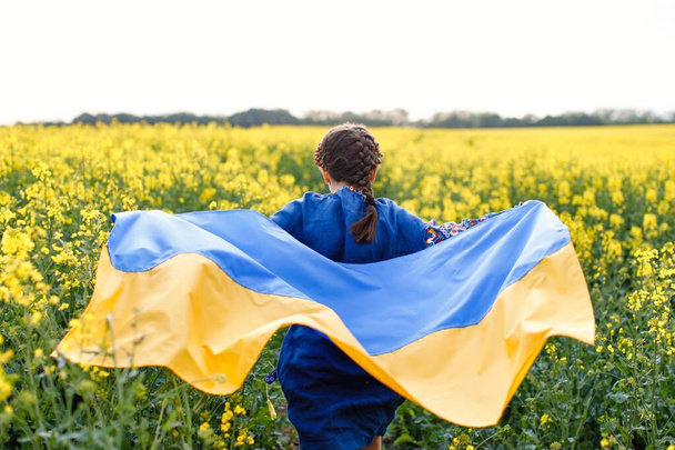 Pray for Ukraine. Child with Ukrainian flag in rapeseed field. A girl in an embroidered shirt runs across the field with the Ukrainian flag in her hands - Valokuva, kuva