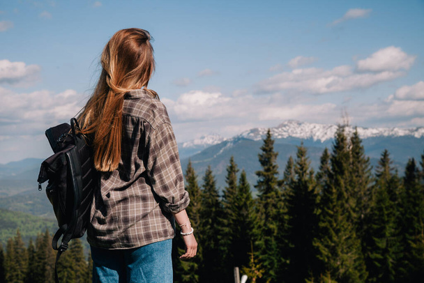 A young, slender girl with loose hair in a plaid shirt and jeans with a black backpack stands against the backdrop of mountains in sunny weather. View from the back. - Fotografie, Obrázek