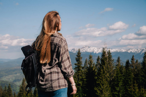 A young, slender girl with loose hair in a plaid shirt and jeans with a black backpack stands against the backdrop of mountains in sunny weather. View from the back. - Foto, Imagen
