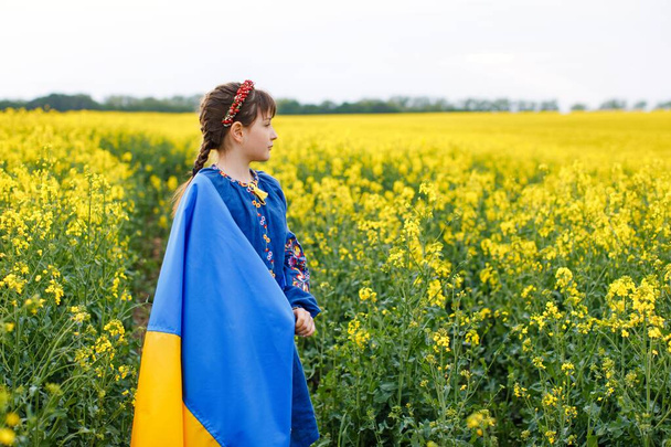 Pray for Ukraine. Child with Ukrainian flag in rapeseed field. Girl holding national flag in her hand. - Фото, зображення