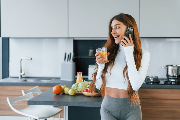 With fresh orange juice. Young european woman is indoors at kitchen indoors with healthy food. - Фото, изображение