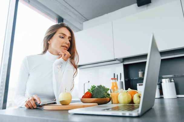 Uses laptop. Young european woman is indoors at kitchen indoors with healthy food. - Valokuva, kuva