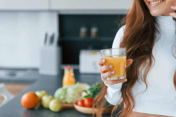 With fresh orange juice. Young european woman is indoors at kitchen indoors with healthy food. - Фото, зображення
