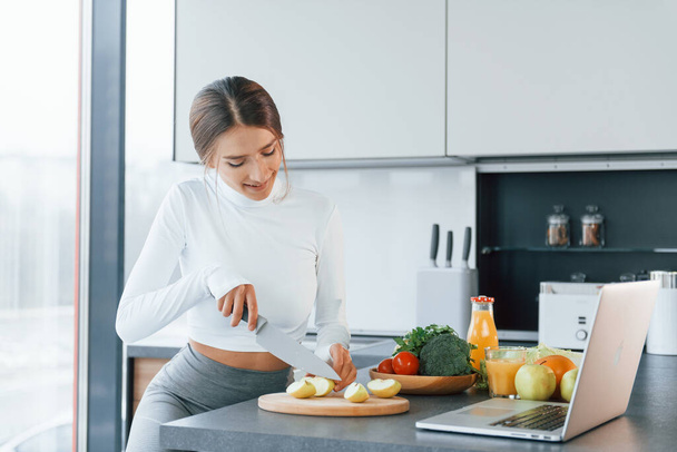 Uses laptop. Young european woman is indoors at kitchen indoors with healthy food. - Fotoğraf, Görsel