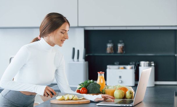 Uses laptop. Young european woman is indoors at kitchen indoors with healthy food. - Foto, Imagem