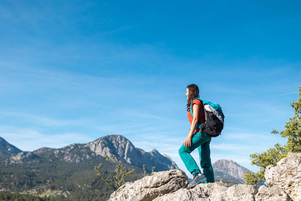 Mladá žena s batohem vyšplhala na vrchol hory a dívá se do údolí, pěší turistika v horách, úspěch při dosahování cíle - Fotografie, Obrázek