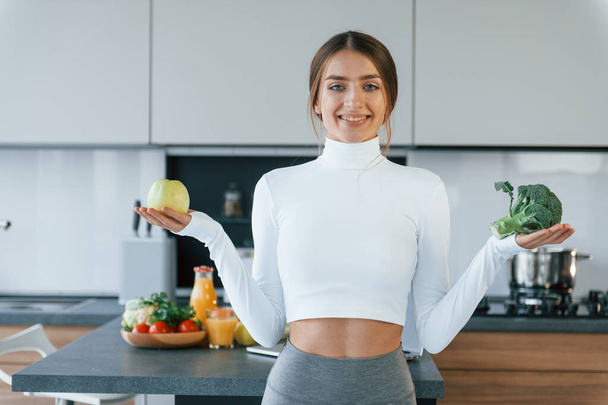 Poses for a camera with vegetables. Young european woman is indoors at kitchen indoors with healthy food. - Photo, image
