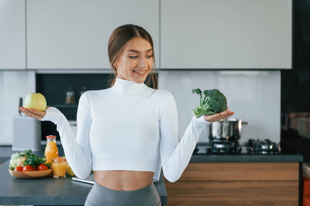 Poses para uma câmera com legumes. Jovem mulher europeia está dentro de casa na cozinha em casa com comida saudável. - Foto, Imagem