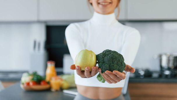 Poses for a camera with vegetables. Young european woman is indoors at kitchen indoors with healthy food. - Foto, Imagen