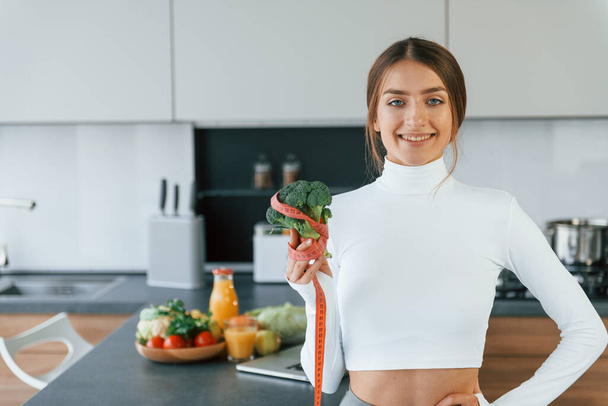 Poses for a camera with vegetables. Young european woman is indoors at kitchen indoors with healthy food. - Photo, image