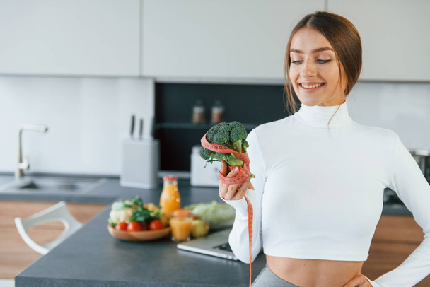 Poses for a camera with vegetables. Young european woman is indoors at kitchen indoors with healthy food. - Фото, изображение