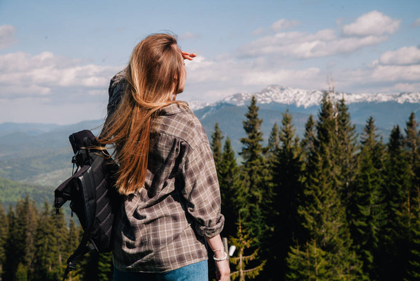 A young, slender girl with loose hair in a plaid shirt and jeans with a black backpack stands against the backdrop of mountains in sunny weather. View from the back. - Photo, image