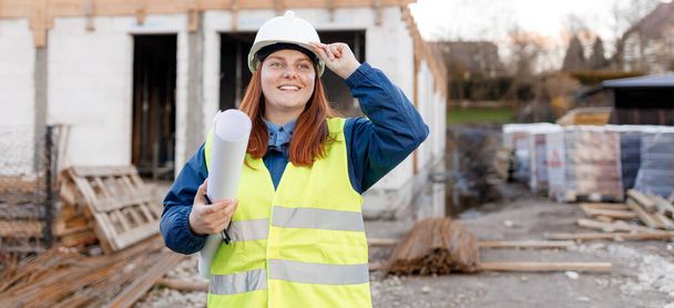 Architect with a blueprints at a construction site. Portrait of successful woman constructor wearing white helmet and safety yellow vest. Woman are planning new project. - Foto, imagen