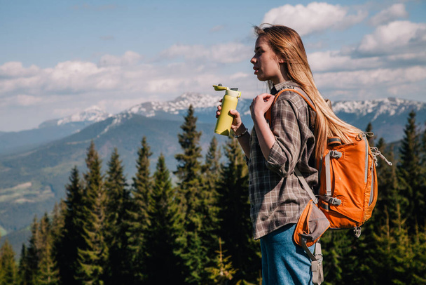 A young, girl in a plaid shirt and jeans with loose hair with an orange backpack drinks tea from a bright thermal cup against the backdrop of the mountains in the Carpathians. view from the back. - 写真・画像