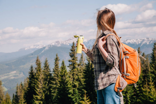 A young, girl in a plaid shirt and jeans with loose hair with an orange backpack drinks tea from a bright thermal cup against the backdrop of the mountains in the Carpathians. view from the back. - Photo, image