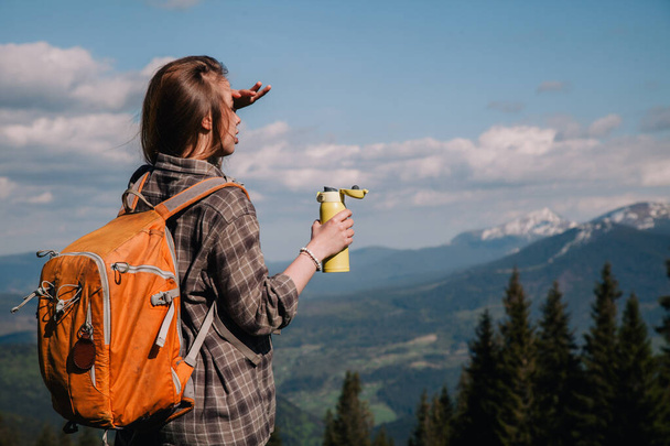 A young, girl in a plaid shirt and jeans with loose hair with an orange backpack drinks tea from a bright thermal cup against the backdrop of the mountains in the Carpathians. view from the back. - Foto, immagini