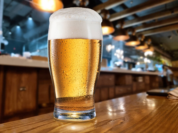 Cooled glass of beer on the wooden table. Blurred bar interior at the background. - Foto, imagen