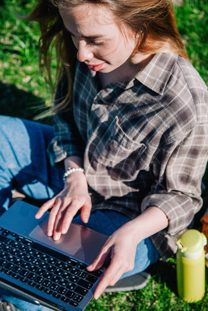 A young, slender girl with loose hair in a plaid shirt and jeans with an orange backpack works on a laptop sitting on green grass against the backdrop of mountains. view from above - Fotografie, Obrázek