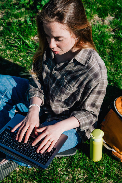 A young, slender girl with loose hair in a plaid shirt and jeans with an orange backpack works on a laptop sitting on green grass against the backdrop of mountains. view from above - Фото, зображення