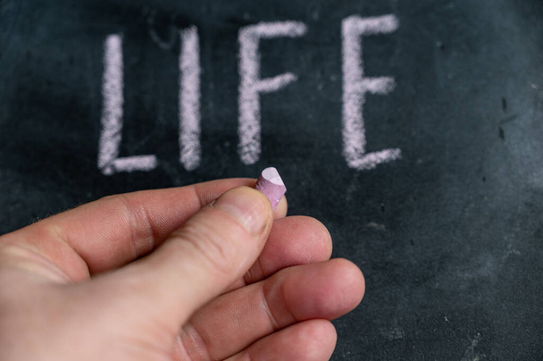 Adult male holding a piece of colored chalk in his hand. The handwritten word LIFE on the black chalkboard. - 写真・画像