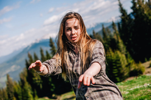 a young, slender girl with loose hair in a plaid shirt and jeans poses for sunny weather in the mountains - 写真・画像