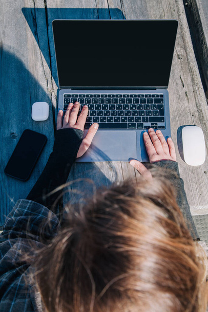 a young, beautiful, slender girl with loose hair in a plaid shirt working on a laptop at an old wooden table in the fresh air. view from above. - Foto, Bild