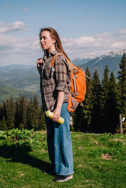 A young, girl in a plaid shirt and jeans with loose hair with an orange backpack drinks tea from a bright thermal cup against the backdrop of the mountains in the Carpathians. view from the back. - Foto, Bild