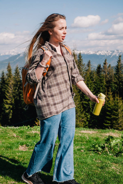 A young, girl in a plaid shirt and jeans with loose hair with an orange backpack drinks tea from a bright thermal cup against the backdrop of the mountains in the Carpathians. view from the back. - Фото, изображение