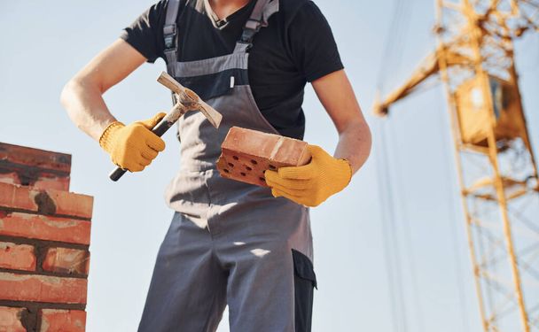 Držet cihlu a používat kladivo. Stavební dělník v uniformě a bezpečnostním vybavení mají práci na stavbě. - Fotografie, Obrázek
