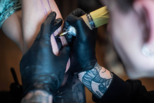 oříznutý záběr tetovacího procesu na ruce v salónu. Profesionální tatér zavádí inkoust do kůže pomocí jehly z tetovacího stroju.Profesionální tatér pracující ve studiu. - Fotografie, Obrázek