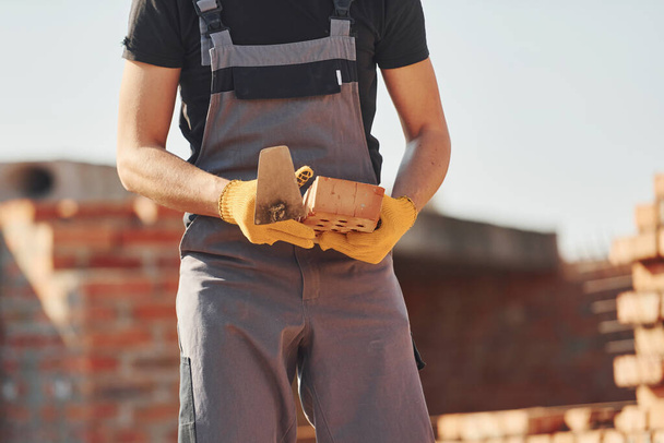 Holds brick in hands. Construction worker in uniform and safety equipment have job on building. - Фото, изображение