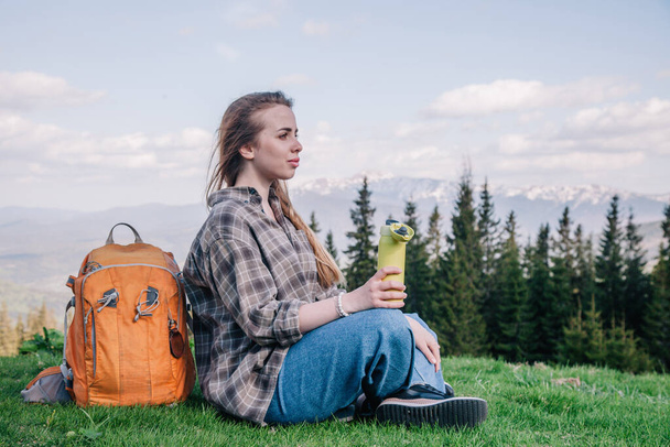 A young, slender girl with loose hair in a plaid shirt and jeans with an orange backpack drinks tea from a thermal mug sitting on the green grass against the backdrop of the mountains. - Photo, image