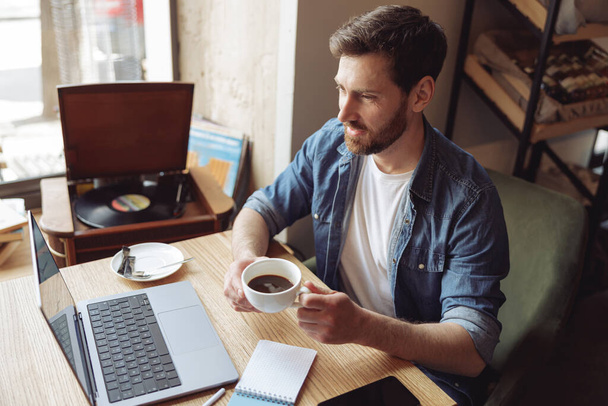Mavi kot gömlekli, kahve içen ve dizüstü bilgisayar ekranına bakan beyaz bir adam.. - Fotoğraf, Görsel