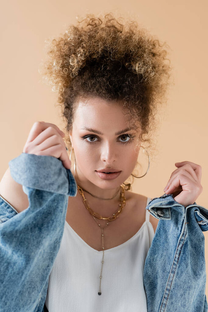 Portrait of stylish model in denim jacket looking at camera isolated on beige  - Foto, Imagen