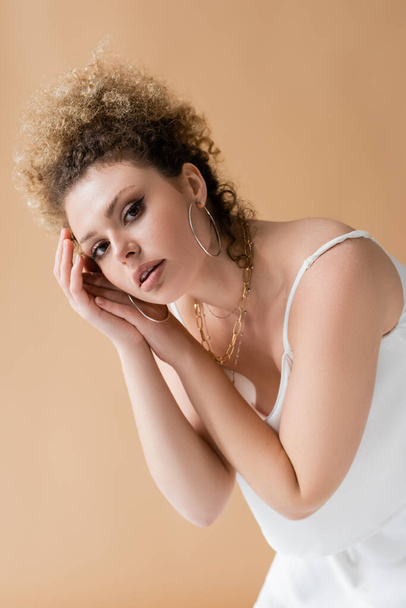 Portrait of curly woman in accessories looking at camera isolated on beige  - Фото, изображение