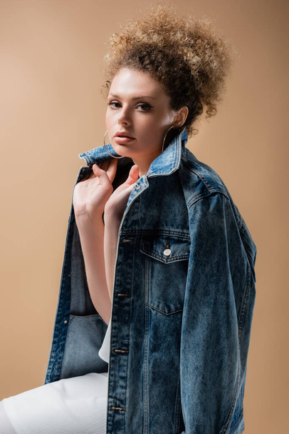 Young woman holding denim jacket and looking at camera isolated on beige  - Фото, изображение