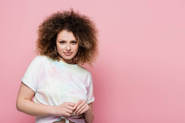 Portrait of smiling curly woman in t-shirt looking at camera isolated on pink  - Foto, Imagen