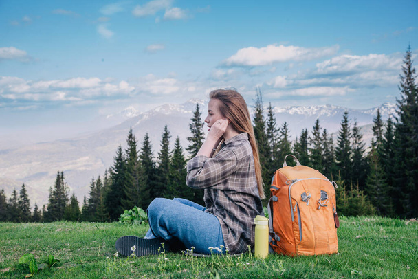 A young, slender girl in a plaid shirt and jeans with an orange backpack meditates sitting on the green grass against the backdrop of mountains. - Fotó, kép