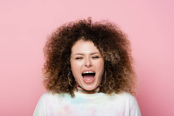 Excited curly woman in t-shirt closing eyes isolated on pink  - 写真・画像