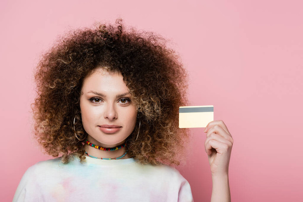 Göndör nő kezében hitelkártya és nézi kamera elszigetelt rózsaszín  - Fotó, kép