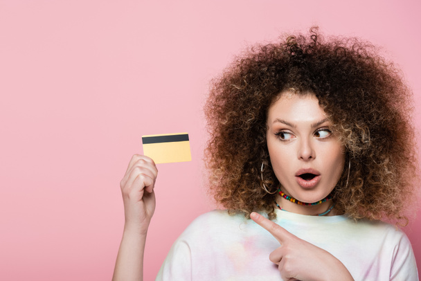 Excited woman pointing at credit card with finger isolated on pink  - Foto, Bild