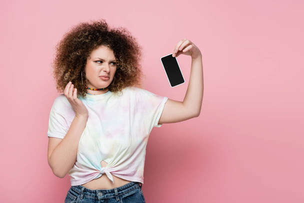 Αηδιαστική νεαρή γυναίκα κρατώντας το κινητό τηλέφωνο με λευκή οθόνη σε ροζ φόντο - Φωτογραφία, εικόνα