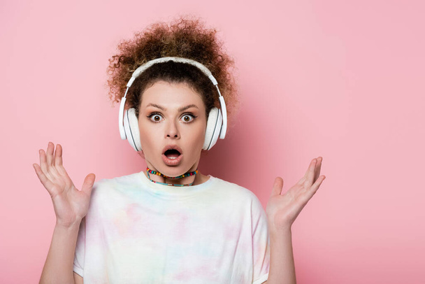 Shocked woman in headphones looking at camera on pink background - Фото, изображение