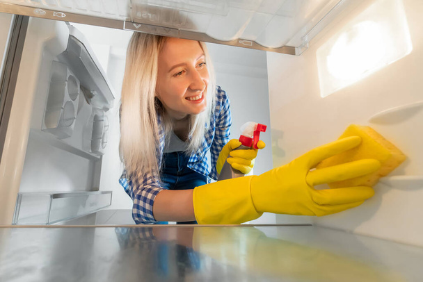 Вид из холодильника. Улыбающаяся женщина в резиновых перчатках и с помощью чистящего средства чистит пустой холодильник губкой. - Фото, изображение