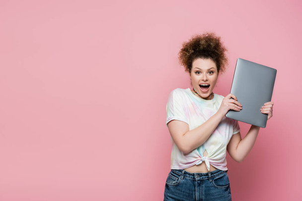 Amazed curly woman holding laptop isolated on pink  - Photo, Image