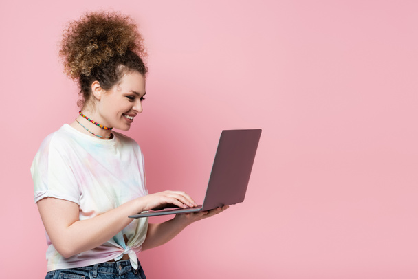 Smiling woman in t-shirt using laptop isolated on pink  - Fotó, kép